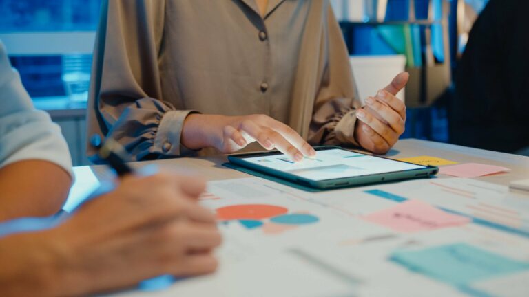A closeup of a table with printouts and a tablet. Two people are sitting at the table, one of them is using the tablet.